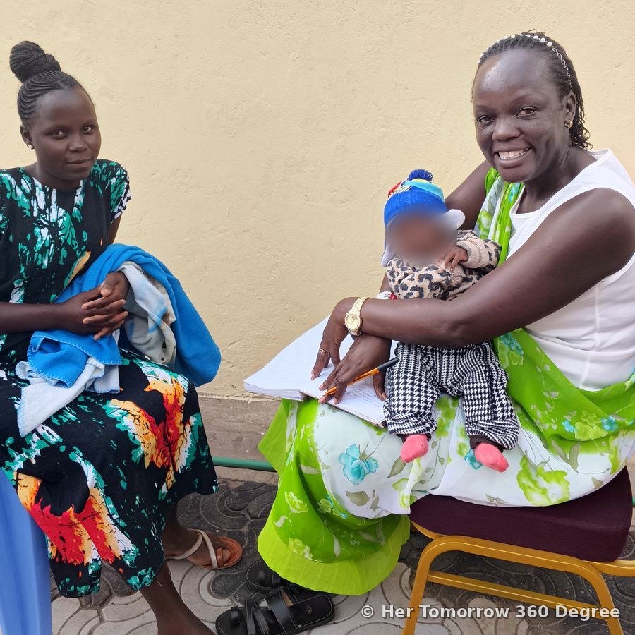 Chairperson Lyma Kivali, smiling, holds a baby while talking with a young mother